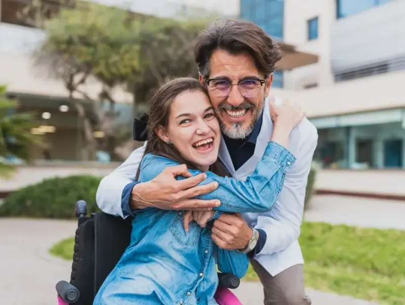 A man embracing a young woman in a wheelchair, both laughing joyfully outdoors