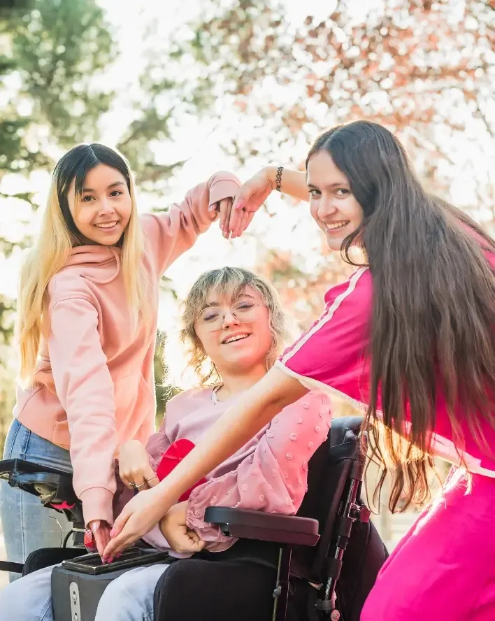 Three young women smiling together outdoors, one in a wheelchair, showing friendship and inclusion